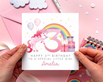 May include: A square birthday greeting card featuring a pink unicorn, a rainbow, balloons, and a large pink number 3. The text reads: Happy 3rd Birthday to a special little girl, Amelia. The design includes stacked gift boxes and yellow stars.