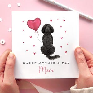 May include: A Mother's Day card featuring a black dog illustration holding a pink heart-shaped balloon. The card reads "Happy Mother's Day Mum" with pink hearts. The card is held against a pink background with decorative elements.