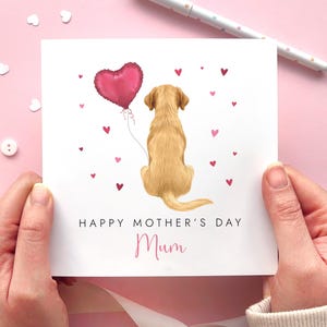 May include: A greeting card for Mother's Day, held by hands, featuring a golden Labrador Retriever holding a pink heart-shaped balloon. The card has the text "HAPPY MOTHER'S DAY" and "Mum" in a script font, with small pink hearts.