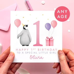 May include: A birthday card with a penguin wearing a party hat and holding a pink balloon. The card has the number 1, two balloons, a gift, and the text "Happy 1st Birthday to a Special Little Girl". The card is held by hands.