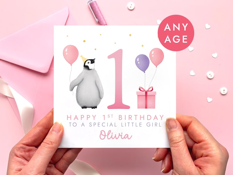 May include: A birthday card with a penguin wearing a party hat and holding a pink balloon. The card has the number 1, two balloons, a gift, and the text "Happy 1st Birthday to a Special Little Girl". The card is held by hands.