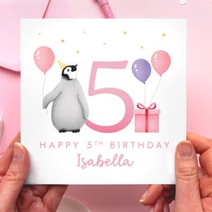 May include: A birthday card with a penguin wearing a party hat, holding a pink balloon. The card features a large pink number 5, two balloons, a gift, and the text "Happy 5th Birthday Isabella."