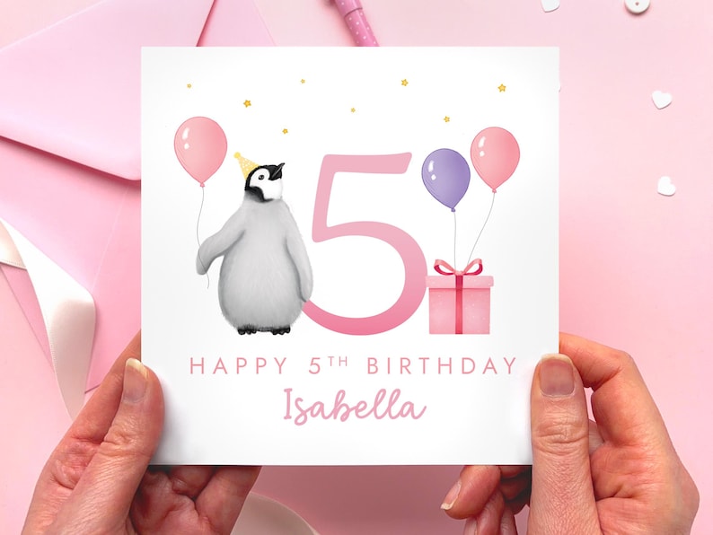 May include: A birthday card with a penguin wearing a party hat, holding a pink balloon. The card features a large pink number 5, two balloons, a gift, and the text "Happy 5th Birthday Isabella."