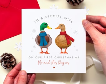 May include: A square greeting card featuring two ducks wearing scarves, one blue and one red, with snowflakes. The text reads: TO A SPECIAL WIFE ON OUR FIRST CHRISTMAS AS Mr and Mrs Rogers, with a small red heart below the names.