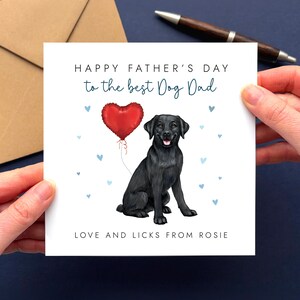 May include: A Father's Day card held by hands, featuring a black Labrador illustration with a red heart balloon. Text reads "Happy Father's Day to the best Dog Dad" and "Love and Licks from Rosie". A brown envelope is in the background.
