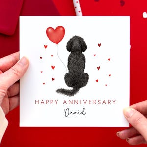 May include: A white greeting card with a black dog illustration holding a red heart balloon. The card reads "HAPPY ANNIVERSARY" and "David" in a handwritten font, with small red hearts scattered around.