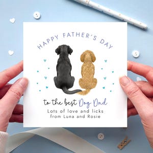 May include: A Father's Day card featuring illustrations of two dogs, a black Labrador and a golden doodle, with the text "HAPPY FATHER'S DAY" and "to the best Dog Dad". The card is held by hands against a light blue background.