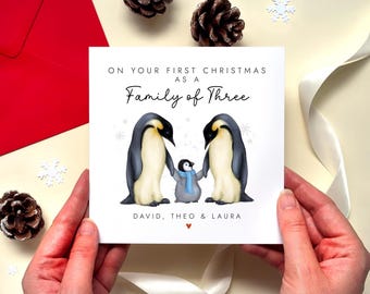 May include: A square greeting card featuring an illustration of two adult penguins and a small penguin chick wearing a blue scarf. The text reads ON YOUR FIRST CHRISTMAS AS A Family of Three and DAVID, THEO & LAURA with a small red heart icon.