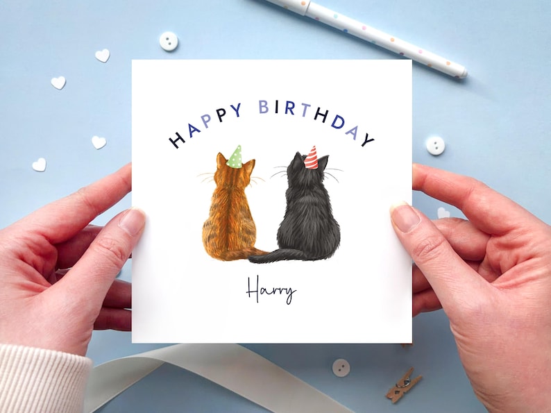 May include: A square birthday card featuring two cats wearing party hats. The text "HAPPY BIRTHDAY" is arched above the cats, with the name "Harry" written below. The cats are a ginger tabby and a black cat.