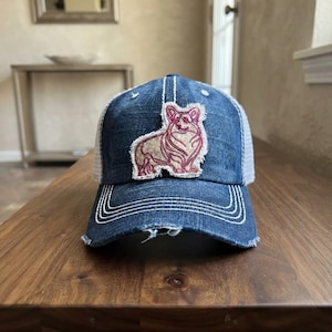 May include: A distressed blue denim baseball cap featuring a white mesh back and a frayed-edge patch on the front. The patch displays an embroidered outline of a Corgi dog in shades of pink and cream with detailed stitching throughout the design.