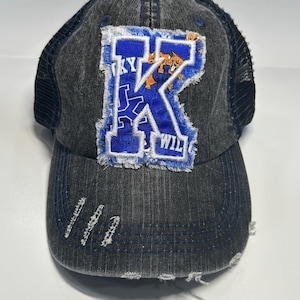 May include: A charcoal grey distressed baseball cap featuring a large, frayed-edge blue patch with a white letter K and a small orange mascot graphic. The cap has a mesh back, a curved brim with frayed detailing, and blue stitching accents.