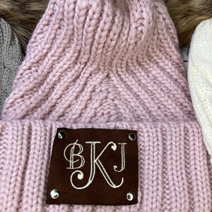 May include: A light pink knitted beanie with a faux fur pom-pom. The hat features a brown leather patch with the embroidered initials "BKJ". The beanie is made of a thick, ribbed knit material.