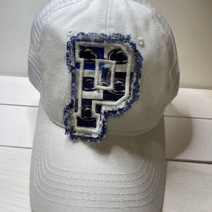 May include: White baseball cap with a mesh back and a curved brim. The cap features a large, distressed blue and white letter "P" patch on the front. The patch has a frayed edge and a patterned design. The cap has a button on top.