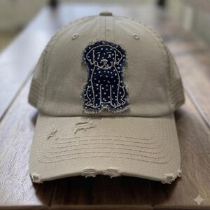 May include: A beige baseball cap with a distressed, frayed brim and front panel. Centered on the front is a dark blue fabric patch featuring a white polka dot pattern and a white embroidered outline of a sitting dog with floppy ears.