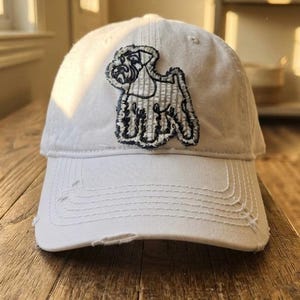 May include: A white baseball cap featuring a dark navy blue embroidered patch of a Schnauzer dog on the front panel. The cap has a distressed, frayed edge along the brim and a structured crown with stitched eyelets for ventilation.