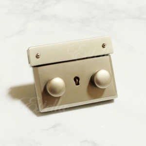 May include: A silver-toned purse clasp with a rectangular body, two rounded knobs, and a keyhole-shaped opening. The clasp has a rectangular top piece with two small screws. The Estella logo is in the bottom right corner.