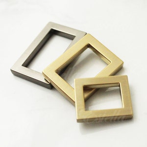 Könnte beinhalten: Drei Metallringe, einer silberfarben und zwei golden, liegen übereinander. Die Ringe sind alle quadratisch mit abgerundeten Kanten.