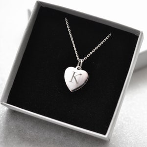 May include: A silver necklace with a heart-shaped pendant engraved with the letter K. The necklace is displayed in a black velvet jewellery box.