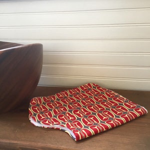 以下が含まれることがあります： 幾何学模様の赤い黄色の布製の鍋つかみ。鍋つかみは半分に折り畳まれ、木製の表面に置かれています。