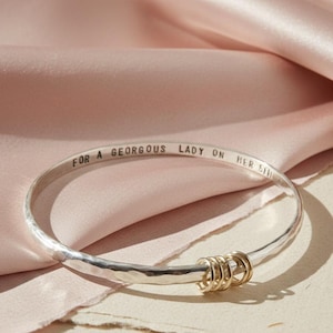 May include: A silver bangle bracelet with a hammered texture and a polished finish. The interior is engraved with the text FOR A GEORGOUS LADY ON HER 5TH. Three small, thin gold-toned rings are threaded onto the bangle, resting at the bottom.