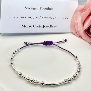 May include: A silver beaded bracelet with a purple adjustable cord. The bracelet features a Morse code design. A card behind the bracelet reads "Stronger Together" and "Morse Code Jewellery".