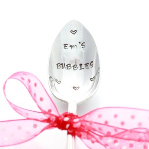 May include: A silver spoon with the words "Em's Bubbles" engraved on it. The spoon is decorated with small hearts and is tied with a pink ribbon with white polka dots.