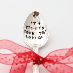 May include: A silver teaspoon with the words "It's time to pour tea Louisa" engraved on it. The spoon is tied with a red ribbon with white polka dots.