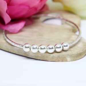 May include: A silver bangle bracelet with five round beads. The bracelet is on a wooden surface.