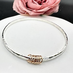 May include: A silver bangle bracelet with a hammered finish and a stack of gold rings attached to the side.