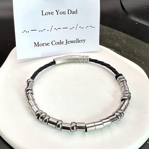 May include: A black leather bracelet with a silver clasp and silver beads that spell out "Love You Dad" in Morse code. The bracelet is on a white surface with a card that reads "Love You Dad" and the Morse code for the phrase. The card also says "Morse Code Jewellery".
