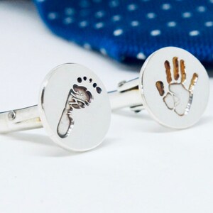 May include: Silver cufflinks featuring a handprint and a footprint design. The cufflinks are round and have a polished finish.