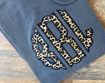 May include: A gray long-sleeved shirt with a large monogram in a leopard print design. The monogram features the letters "SRB" in a decorative font. The shirt also has the text "Sew Southern Monograms" in a circular design.