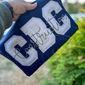 May include: A navy blue sweatshirt with white felt letters that spell "CRC" and the word "Construction" embroidered in gray thread.