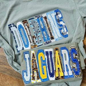 May include: Two grey t-shirts with the words "Dolphins" and "Jaguars" spelled out in colourful patchwork letters. The letters are made of different fabrics and patterns, including blue, yellow, white and black.