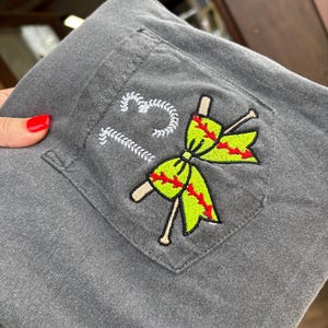 Op de afbeelding: Een grijs t-shirt met een zak met een geborduurd softball-strikje met gekruiste knuppels en het nummer "12" erboven.