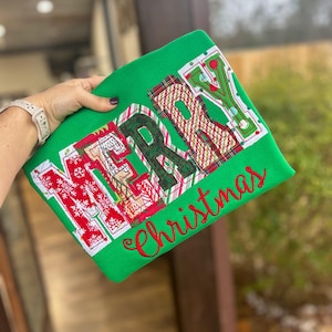 Puede incluir: Camiseta verde con un diseño de "Feliz Navidad" en rojo, blanco y verde. Las letras están hechas de tela de patchwork con una variedad de patrones.