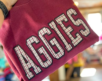 May include: Burgundy sweatshirt with a large "Aggies" graphic in white and gray plaid.