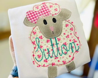 May include: White t-shirt with a pink and white floral applique of a sheep with a pink and white gingham bow. The sheep has the name "Sutton" embroidered on it in teal thread.