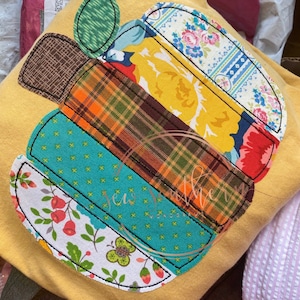 May include: A yellow fabric with a patchwork pumpkin design. The pumpkin is made of different fabrics with floral, plaid and geometric patterns. The pumpkin is outlined in black thread.