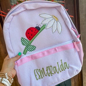May include: A light purple and white seersucker backpack featuring an embroidered red ladybug on a green stem with a white daisy. The front pocket has a horizontal pink stripe and the name Esmeralda embroidered in green cursive script.