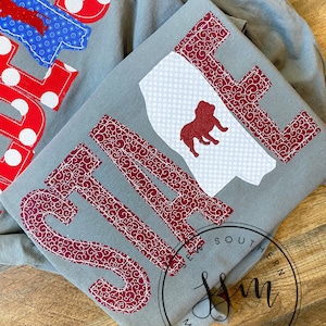 Puede incluir: Una sudadera gris con una aplicación granate de la palabra "STATE" con un contorno blanco y un fondo blanco con una silueta de bulldog granate. La sudadera está doblada por la mitad y la parte inferior de la sudadera tiene un círculo negro con el texto "Sew Southern Monograms".