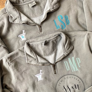 Puede incluir: Dos sudaderas grises con monogramas bordados. La sudadera de arriba tiene el monograma "LSB" en azul turquesa. La sudadera de abajo tiene el monograma "DMP" en azul turquesa y un símbolo blanco de mortero y maja. La sudadera de abajo también tiene un círculo blanco con el texto "Sew Southern SSM Monograms".