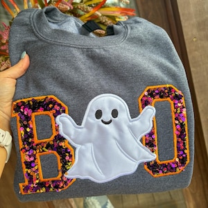 May include: A dark heather gray crewneck sweatshirt featuring the word BOO appliqued on the front. The letters B and O are filled with purple and orange sequins and outlined in orange fabric. A white ghost with a black face sits in the middle.