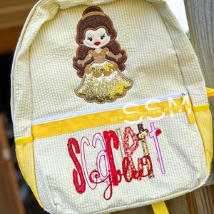 May include: Yellow and white striped fabric backpack with a yellow zipper and yellow straps. The backpack has a yellow and white striped fabric pocket on the front with a red, pink, and white embroidered name "Scarlett". The top of the backpack has a brown haired, yellow dress, and gold glitter embroidered character that resembles Belle from the Disney movie Beauty and the Beast.