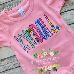 May include: Pink baby onesie with the name "Kyndall" spelled out in colorful patterned fabric. The onesie has a floral design and the text "Sew Southern Monograms" at the bottom.