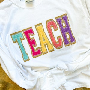 May include: White long-sleeve t-shirt with the word "TEACH" in colorful, fuzzy letters with gold glitter accents.