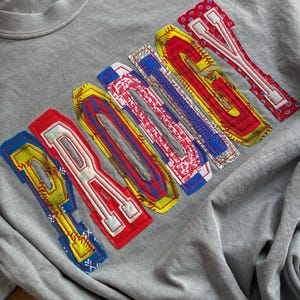 Op de afbeelding: Grijze crewneck sweatshirt met het woord "PARADISE" op de borst. De letters zijn in een kleurrijk patchwork-ontwerp, omlijnd in wit, met patronen en kleuren waaronder rood, geel, blauw en wit.