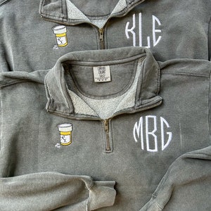 Puede incluir: Dos sudaderas grises con cierre de cremallera. Una sudadera tiene un monograma "KJE" y la otra tiene un monograma "MBG". Ambas sudaderas tienen una pequeña imagen bordada de un frasco de pastillas con una etiqueta de receta.