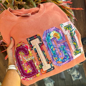 May include: A peach-coloured crewneck sweatshirt featuring the word GIGI in large, fabric-appliqued varsity letters. Each letter is crafted from a different floral and patterned fabric with visible stitching around the edges of the letters.