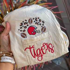 Op de afbeelding: Een crèmekleurig T-shirt met een geborduurd ontwerp boven de borstzak. Het ontwerp bevat een rode footballhelm, drie bruine footballs en luipaardprintvlekken. Het woord "tigers" is in rode schrijfletters op de zak geborduurd.